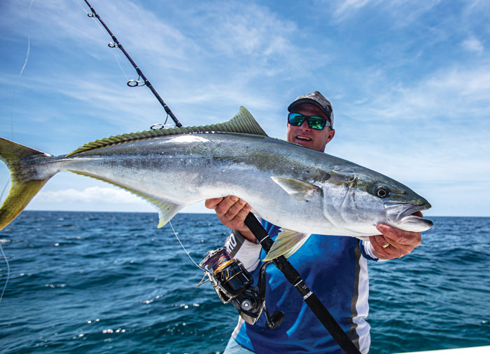 Man with fishing rod holding Yellowtail Kingfish with blue ocean behind